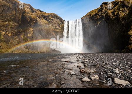 Inquadratura a basso angolo del maestoso paesaggio della cascata di Skógafoss, con un cielo blu chiaro e un bellissimo arcobaleno, vicino alla Route 1 / Ring Road, Islanda Foto Stock
