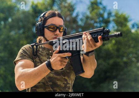 Primo piano ritratto di un uomo maschile muscoloso militare-come nero-capelli bearded uomo in moro t-shirt guardando da vicino il suo fucile nero. Alberi sfocati sullo sfondo. Scatto medio per esterni. Foto di alta qualità Foto Stock