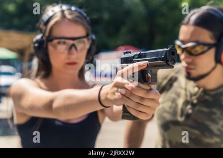 Un giorno in un campo di tiro. Ragazza adulta caucasica fiduciosa che punta al bersaglio con una pistola nera. Occhiali e cuffie di protezione. Lezione privata di tiro. Foto di alta qualità Foto Stock