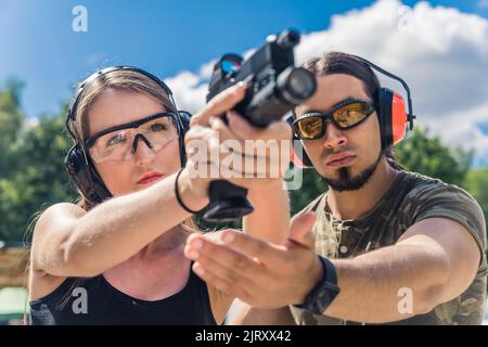 Primo piano all'aperto di una donna caucasica focalizzata in cuffie protettive e occhiali che puntano al bersaglio. Istruttore di tiro muscolare che corregge la sua postura. Foto di alta qualità Foto Stock