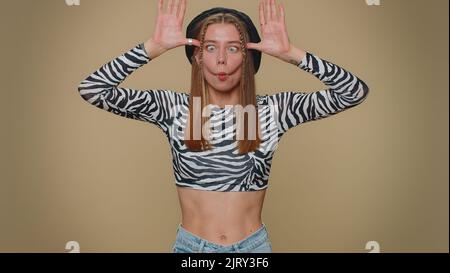 Bella donna piuttosto divertente in crop top facendo divertenti espressioni facciali sciocco e grimacing, imbrogliando intorno, mostrando la lingua. Ragazza donna elegante adulto isolato da solo su sfondo beige studio al coperto Foto Stock