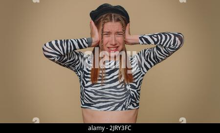 Non vuoi ascoltare e ascoltare. Frustrata irritata giovane donna piuttosto irritata coprendo le orecchie, gesturando no, evitando consiglio ignorando sgradevole rumore voci forte. Ragazza elegante adulto su sfondo beige Foto Stock