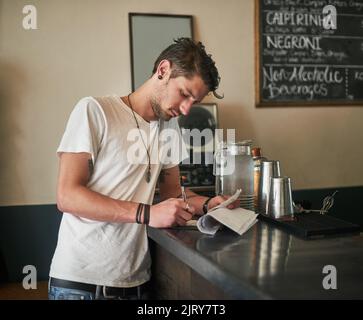 Un altro giorno nella vita di un barkeep. Un barman giovane e focalizzato che prende appunti sulle ricevute dietro il bar. Foto Stock