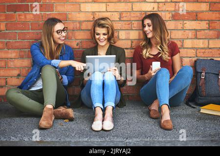 Raffreddare insieme nel campus. Tre sorridenti studenti universitari seduti insieme fuori dal campus utilizzando un tablet digitale. Foto Stock