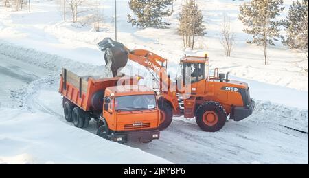 Dicembre 23, 2021. Regione di Kemerovo, Russia. Il trattore arancione grande pulisce la neve dalla strada e la carica nel carrello. Pulizia e pulizia della strada Foto Stock