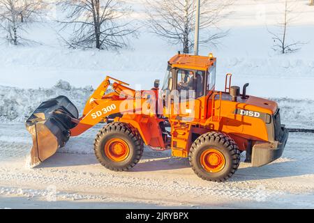 Dicembre 23, 2021. Regione di Kemerovo, Russia. Il trattore arancione grande pulisce la neve dalla strada e la carica nel carrello. Pulizia e pulizia della strada Foto Stock