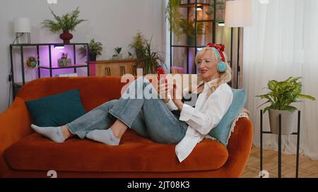 Felice rilassato donna anziana gioita in cuffie wireless ballare sul divano a casa scegliendo l'ascolto di musica disco rock n roll preferito energica in smartphone divertente. Persone attività weekend Foto Stock