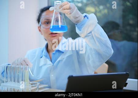 Concentrarsi su un pallone a fondo piatto in mano a un chimico che ispeziona la reazione chimica che avviene in soluzione blu Foto Stock