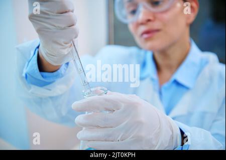 Concentrarsi sulla pipetta di vetro da laboratorio in mani guantate di una farmacologa femminile sfocata che gocciola pochi reagenti nel matraccio Foto Stock