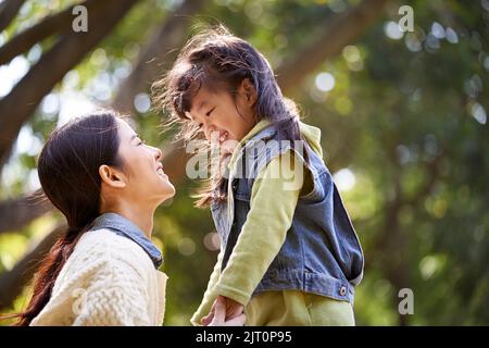 giovane madre asiatica seduta sull'erba nel parco avendo una conversazione piacevole con la figlia carina Foto Stock