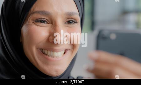 Headshot giovane bella felice donna araba in hijab sorridente con denti bianchi sani guardando lo schermo dello smartphone gioire di buone notizie guardando divertente Foto Stock