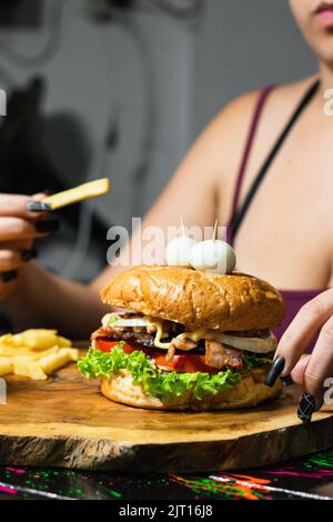 primo piano di una ragazza latina con il suo hamburger e patatine servite sul tavolo di legno, pronto per iniziare a mangiare. donna con una patata in mano circa per mettere Foto Stock