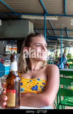 Bella e felice donna bionda bere birra in un ristorante in America Latina. Indossa un bikini giallo Foto Stock