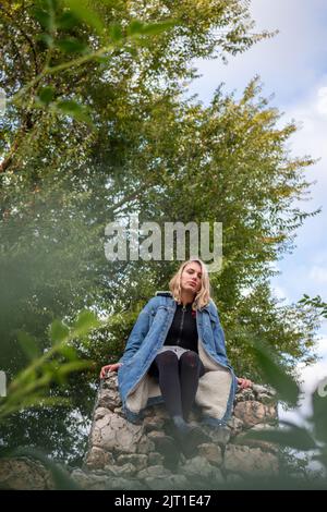 Bella donna bionda seduta su una roccia in un parco verde. È triste Foto Stock