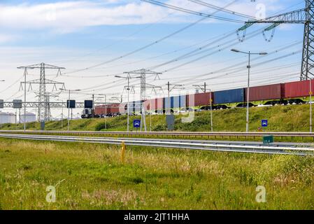 Treno cargo carico di container che percorrono un'autostrada in una zona portuale in una chiara giornata estiva. Piloni elettrici e serbatoi carburante sono in background. Foto Stock