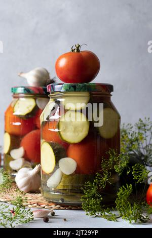 Zucchine sottaceto con pomodori in due vasetti di vetro su fondo grigio chiaro con ingredienti freschi, formato verticale Foto Stock