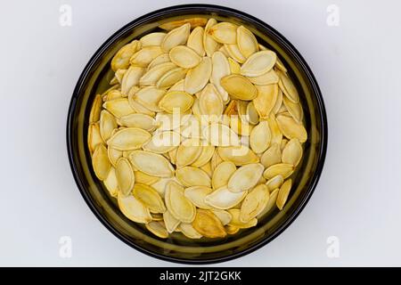 Semi di zucca essiccati in bucce in una ciotola nera su fondo bianco. Isolato. Semi di zucca gialli in un piatto. Seme ovale piatto di zucca. Cibo sano. Foto Stock