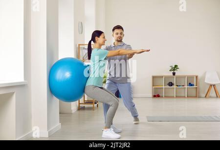 Fisioterapista aiutare il paziente giovane fare squats della parete con la sfera di idoneità per liberarsi di mal di schiena Foto Stock