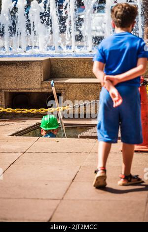 Ragazzo bianco che guarda l'operaio di manutenzione delle fontane che esce dal sottosuolo a Savassi a Belo Horizonte, Brasile. Foto Stock