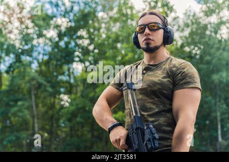 Sparo medio all'aperto di un uomo muscoloso caucasico dall'aspetto militare in una t-shirt moro che guarda lontano dal suo bersaglio e che tiene un fucile nero con una loupe da guardare attraverso. Foto di alta qualità Foto Stock