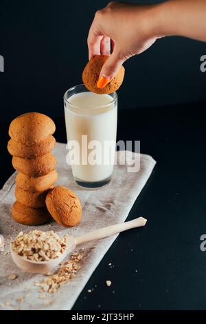 Biscotti di farina d'avena disposti in una pila, la mano di una donna bagna i biscotti in un bicchiere di latte. Sfondo nero. Primo piano del cibo Foto Stock