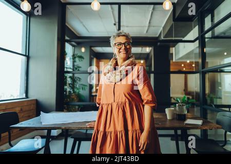 Felice donna d'affari matura sorridente alla macchina fotografica mentre si trova in un ufficio. Allegra donna d'affari matura in piedi nella sala del consiglio di un lavoro moderno Foto Stock