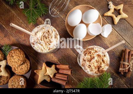 Melanzana con panna montata in bicchieri, uova, frusta, cannella, biscotti natalizi su vecchio tavolo di legno. Il tradizionale drink di Natale è la cucina a partire da uova e mi Foto Stock