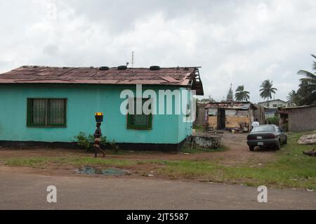 Vista sulla strada e piccola casa verde, Sinkor, Monrovia, Liberia. Signora che porta i secchi sulla sua testa. Foto Stock