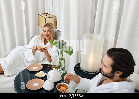 Coppia durante un weekend romantico. Coppia giovane amorevole in accappatoi bianchi che trascorrono del tempo al centro benessere e gustando il tè curativo Foto Stock