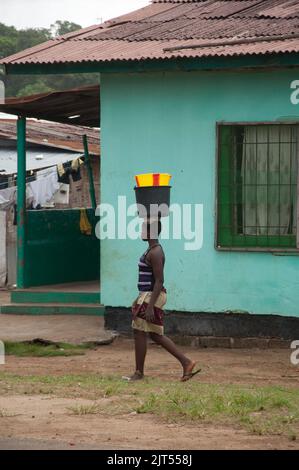 Vista sulla strada e piccola casa verde, Sinkor, Monrovia, Liberia. Donna che porta secchi sulla sua testa. Foto Stock