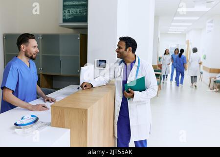 Medico indiano che parla con un'infermiera maschile vicino al banco della reception presso un moderno ospedale durante la giornata lavorativa. Vista della clinica medica della hall con i pazienti e la p Foto Stock