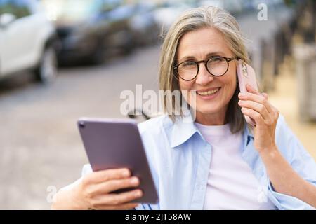 Leggere gli aggiornamenti sul tablet digitale matura donna capelli grigio parlare al telefono seduto sulla panchina per le strade della vecchia città europea. Donna matura che risponde alla chiamata all'aperto. Foto Stock