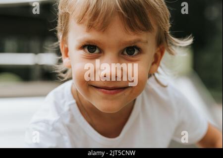 ritratto del volto di un ragazzino carino felice e sorridente di cinque anni con grandi occhi e lunghi capelli biondi in una t-shirt bianca. generation z ch Foto Stock