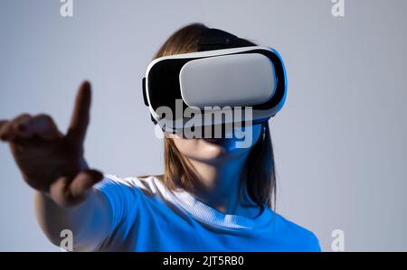 Giovane bruna donna in t-shirt bianca con visore per la realtà virtuale che tocca l'aria durante la realtà virtuale in un'esperienza metaversa. Concetto di tecnologia metaverse Foto Stock