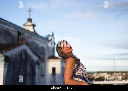 Una donna sul portico della sua casa in abiti leggeri guardando la strada contro il cielo e la chiesa sullo sfondo. Città di Valenzia, Bahia, Brasile. Foto Stock