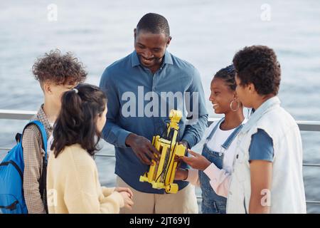 Ritratto di un insegnante sorridente che mostra il modello del robot a un gruppo di bambini in piedi in cerchio durante la lezione di ingegneria all'aperto Foto Stock