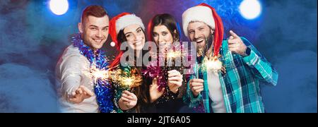 Un gruppo di amici giovani adulti allegri a una festa con cappelli di babbo natale e brandeli con gli scintillanti in mano, ridendo e guardando la fotocamera su Foto Stock