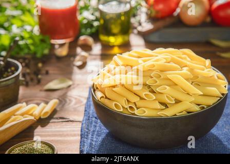Recipiente per pasta di penne crude con ingredienti freschi su fondo rustico in legno. Foto Stock