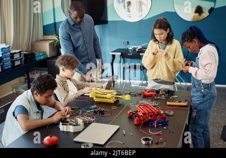 Un gruppo eterogeneo di bambini adolescenti che costruiscono robot insieme mentre si godono la classe di ingegneria nella scuola moderna Foto Stock