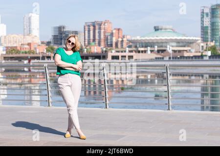 Un ritratto completo di una ragazza felice in una blusa verde, pantaloni e occhiali da sole sullo sfondo di una città sfocata. Una ragazza cammina per la strada Foto Stock