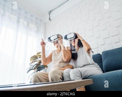 Giovane donna asiatica, figlia felice e sua madre, vecchia donna anziana indossando occhiali VR, godersi il gioco 3D online insieme in stanza bianca. Mamma e figlia Foto Stock