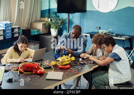 Ritratto di insegnanti neri maschi costruzione robot con diversi gruppi di bambini in classe di ingegneria presso la scuola moderna, copia spazio Foto Stock