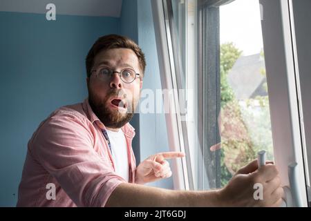 L'uomo scioccato con una strana espressione sul viso si erge vicino ad una finestra aperta e punta il dito nel cortile di sorpresa. Foto Stock