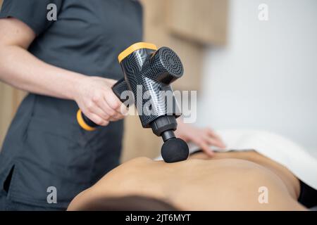 La donna massaggia la schiena del paziente con un massaggiatore a percussione Foto Stock