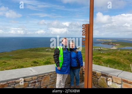 Ritratto di coppia di turisti adulti sorridenti al punto di osservazione di Sky Road contro l'oceano e il cielo blu, la campagna irlandese e lo skyline sullo sfondo, soleggiato s Foto Stock