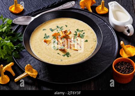 Kanttarellikeitto, zuppa finnica di Chanterelle in ciotola nera su tavolo di legno scuro con cucchiaio, funghi Chanterelle freschi sullo sfondo, vista orizzontale Foto Stock