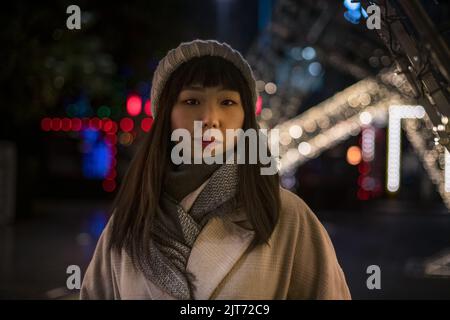 Una giovane bella donna coreana alla moda in un cappello e un cappotto cammina per le strade natalizie della città in serata. Foto Stock