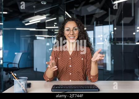 Webcam vista, felice donna d'affari ispanica sorridente e guardando la fotocamera, videochiamata con i colleghi, riunione remota, lavoratrice in ufficio moderno indossando occhiali e capelli ricci Foto Stock