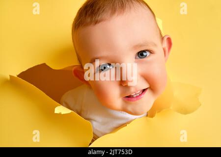 Un bambino felice guarda fuori da un buco nello studio sfondo giallo. Sorridendo il bambino sbirciando attraverso uno sfondo di carta strappato, spazio di copia. Età dei bambini un anno Foto Stock