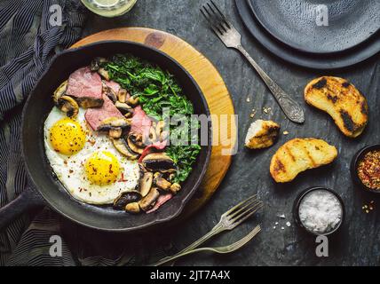 Padella di uova con pancetta, funghi e spinaci serviti con pane e spezie in crosta su fondo scuro. Vista dall'alto, primo piano Foto Stock